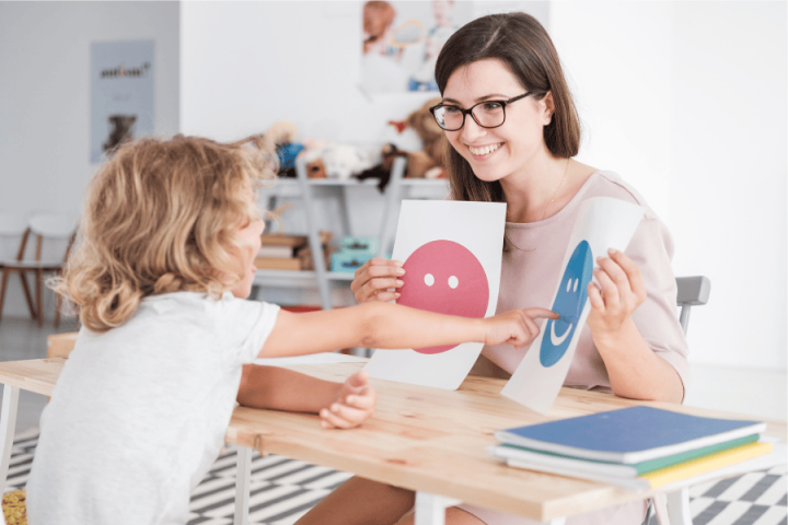 Teacher holding images of happy and sad faces sits in front of a child.